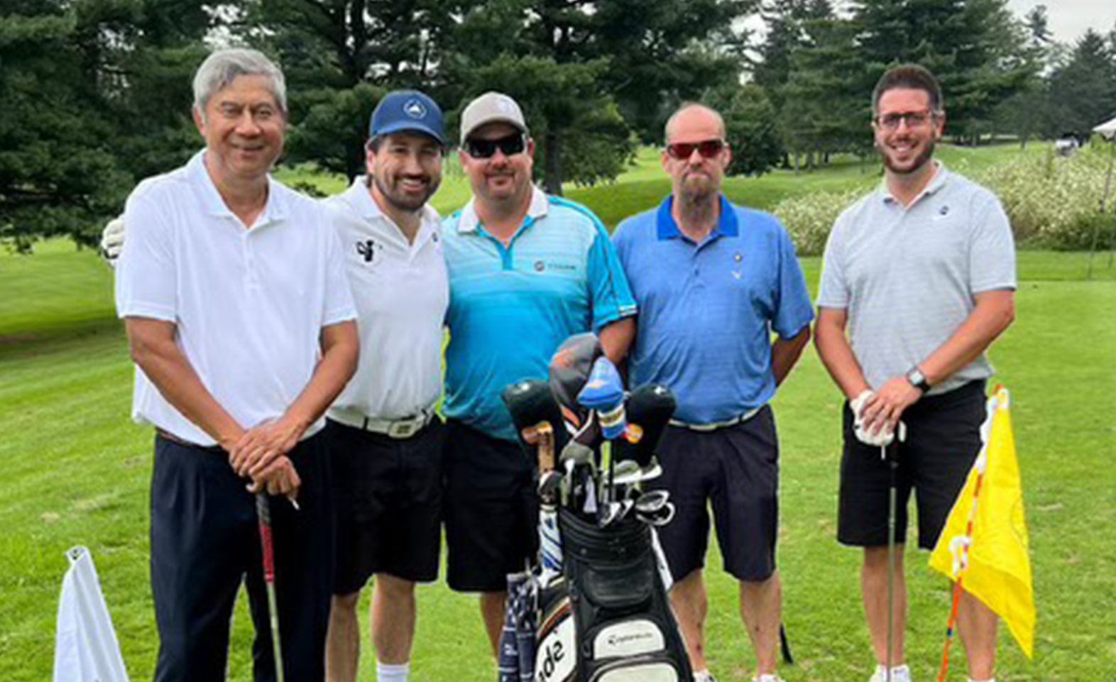 Group of five men posing together on a golf course, with golf clubs and flags in the foreground.
